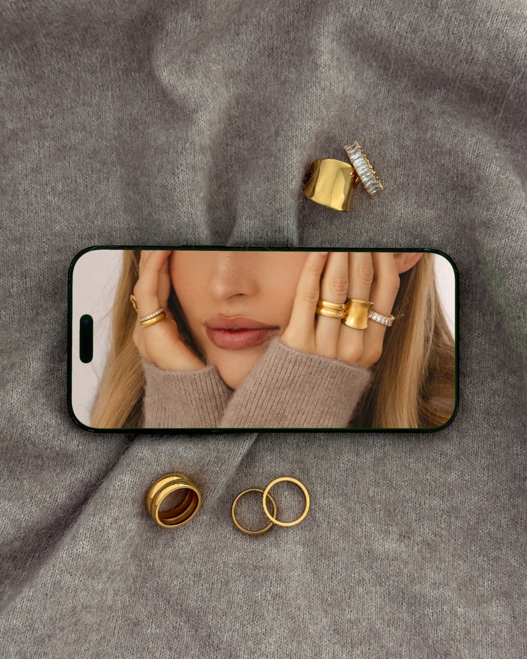 Teléfono móvil mostrando el rostro de una mujer con anillos de oro, rodeado de joyas sobre un fondo de tela gris.